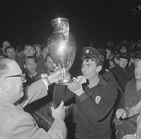 A smiling male goalkeeper is presented with the European Cup trophy while surrounded by a crowd.