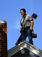 A caucasian man of about forty stands on the ridge of a modern house next to a red brick chimney. He is in jeans and a polo shirt and wears leather safety gloves.On his back is a standard chimney sweeping brush and poles.