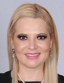 An image of a smiling woman wearing makeup with long blonde colored hair, black shirt, golden earrings and a white necklace.