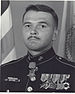 A black and white image of Kellogg from the chest up wearing his military dress blue uniform with medals. There is an American flag and a Marine Corps flag behind him and his Medal of Honor is around his neck.