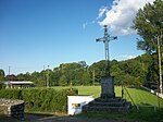 Monument avec une croix en fer forgé. Au centre est représenté le Sacré-Coeur de Jésus, au sommet est placé un coq. En arrière plan, le stade de football