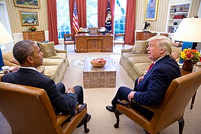 Barack Obama sits in the left Foreground while Donald Trump sits to the right with the ornate Resolute desk center in the background.