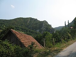 A picture of a lush cliff in the distance. A house from the village of Solotuša with a red roof sits below the path the image is taken from.