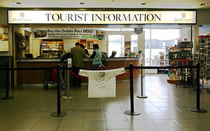 A white t-shirt showing the Wikipedia logo and arious signatures hangs over the queue barrier at a Dublin Tourism tourist information booth. A large sign in the background says, "Buy the Dublin pass here!"