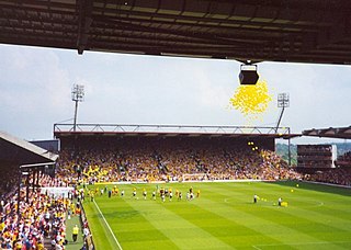 In the background and to the left are two large stands, each of which seem capable of holding thousands of people. In the foreground is a well maintained grass pitch. Yellow balloons can be seen in the sky.