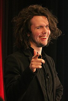 William Pugh, pointing at the camera and smiling. He has long, curly black hair.