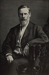 Portrait photographique d'un homme portant lunettes et barbe qui se tient assis sur une chaise.