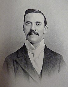 A vintage photograph of a man with neatly combed hair and a moustache, wearing a dark suit, a high-collared shirt, and a cravat, looking directly at the camera with a slight smile.
