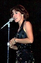 A dark-haired young woman in a black dress singing into a microphone