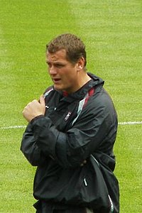 A man wearing a waterproof jacket stands on a football pitch with his arms folded