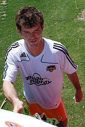 A young man with dark hair stands on a grassy surface. He is wearing a white T-shirt with black trim and orange shorts, and is smiling slightly.