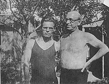 Two middle-aged man pose for an informal photo under some trees. Their arms are behind one another's backs. One man is wearing a t-shirt and the other is bare-chested