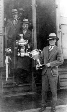 Photographie en noir et blanc de trois hommes descendant d'un train, deux d'entre eux tenant une coupe dans la main