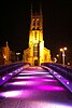 A purple-lighted bridge leading to a yellow-illuminated tower