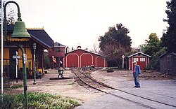 An elderly man standing next to railroad track in the foreground with an old-fashioned railroad depot building on the opposite side. A locomotive shed and water tower are located where the railroad track ends in the background.