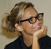 A blonde woman wearing a black shirt and eyeglasses smiles and looks off-camera, holding her chin in her hand.