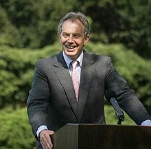 A smiling man with grey hearing wearing a dark suit, standing behind a podium