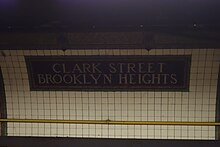 A dark tile mosaic on the curved wall of the station, reading "Clark Street Brooklyn Heights". The rest of the wall is clad with white tile.