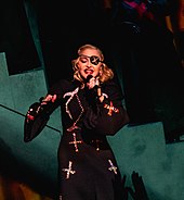 Madonna singing onstage in front of a greenish backdrop, while wearing black costumes and an eye-patch.