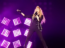 A blonde woman standing in front of purple lights and dressed in black sings to a microphone.