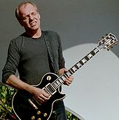 A man in a gray shirt with his eyes closed and a black guitar strapped around his neck.