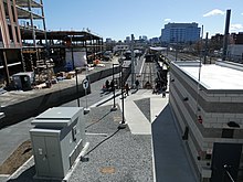 A light rail station in an urban area. A small cinderblock building is at right; a tall building is under construction at left.