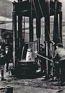 Workers stand around a large forge, where a shell burns red-hot.