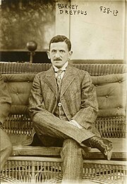 Black and white image of a man with a mustache, wearing a formal, Edwardian era suit and sitting with his legs crossed.