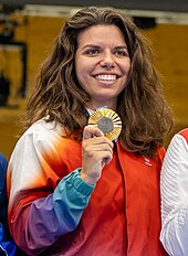 Athlète féminine souriante et montrant sa médaille d'or sur un podium.