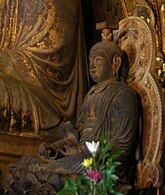 Three-quarter view of cross-legged seated statue placed on a throne. Both arms rest on his legs. The hair is sculpted with a top knot. Color photograph.