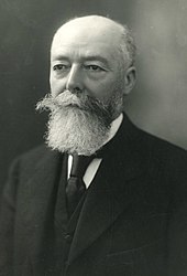 Portrait photographique d'un homme aux cheveux et à la barbe grisonnants, vêtu d’une chemise blanche, d’un veston, d'une veste et d'une cravate foncés
