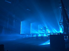 Porter Robinson performing on stage, standing behind a table with equipment, raising his hands. Blue beams of light radiate from the stage.