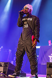 Artist performing onstage in a black jumpsuit reading "nerves of till", pink wig, white plastic sunglasses, and Converse-type sneakers.
