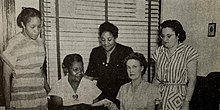 A photograph of three black woman and two white women standing and seated in front of a window covered by a Venetian blind