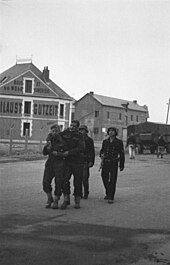 Two wounded commandos escorted by two armed German naval personnel. A large building is in the background