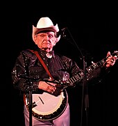 An older man wearing a white cowboy hat and a black dress shirt, standing behind a microphone stand and holding a banjo.