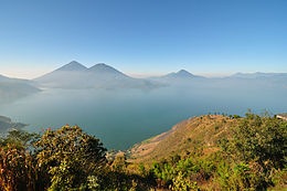 Vue depuis les collines d'un large lac baigné dans la brume. Le lac se trouve aux pieds de plusieurs volcans.