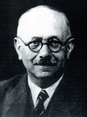 Black-and-white photo of an older man with round glasses and dressed formally in a dark suit, white shirt, and tie