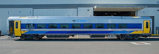 A stainless steel passenger rail car with yellow doors, a dark blue stripe along the windows, and a multi-color stripe along the bottom with yellow plus several shades of blue