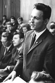 Inside a large room, a short dark-haired man in a suit stands up as he speaks to someone. Behind him, other people dressed in formal attire are all seated.