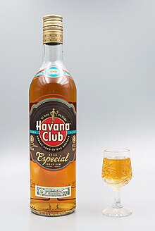 Photo d'une bouteille en verre transparent, rempli de rhum à la couleur ambrée. La bouteille est quasiment remplie, elle a trois étiquettes dont la principale porte le logo de Havana Club. A côté de la bouteille, un petit verre est rempli de rhum.