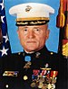 Head and shoulders of a white man wearing a white peaked cap and a dark military jacket with ribbon bars on the right breast, two rows of medals hanging from ribbons on the left breast, and a star-shaped medal hanging from a light-blue ribbon at the neck. Behind him are an American flag and a red and yellow flag.