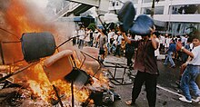 A man wearing a buttoned shirt, pants, and flip-flops throws an office chair into a burning pile of other chairs in the middle of a city street. Behind him, several dozen people gather in front of a building with broken windows.