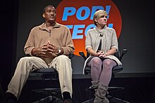 Ronald Cotton (left) and Jennifer Thomson-Cannino (right) sitting in two rolling office chairs in front of large red and blue Pop!Tech logo.