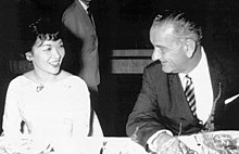 A middle-aged lady wearing a light-coloured dress and with short hair, fluffy at the front, sits at a dinner table smiling. To the right is a taller, older man in a dark suit, striped tie and light shirt who is turning his head to the left, talking to her. A man in a suit is visible, standing in the background.
