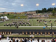 This image shows the North Augusta High School football team running onto a football field through a tunnel. The band can be seen performing in concert arch and there are students running with flags ahead of the team.