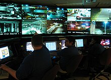 Photo of a security operations center. Contains 5 computers on tables, 10 televisions on the wall, and 2 cybersecurity personnel.