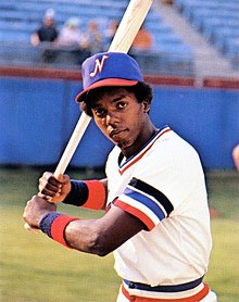 A man wearing a white baseball uniform with a blue cap poses holding a baseball bat as if ready to swing at a ball.