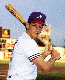 A man wearing a white baseball uniform with a blue cap poses holding a baseball bat with both hands as if ready to swing.