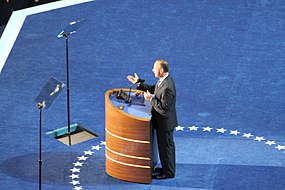 (A) The first three teleprompters: A monitor screen partly embedded in the lectern's desktop displays the scrolling text of the speech in synchrony with two screens embedded in the podium floor. They are on either side of the speaker, reflected by the angled glass teleprompters above them. Click this image and the ones below to see the four-teleprompter system more clearly.